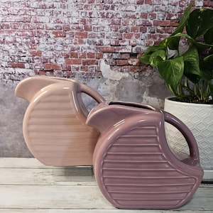 May include: Two ceramic pitchers with a ribbed design. The pitcher on the left is a light pink color, while the pitcher on the right is a darker shade of pink.