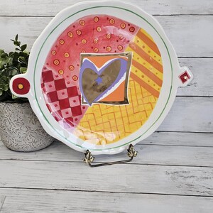 May include: A decorative ceramic plate with a colorful, abstract design. The plate features a heart-shaped design in the center, surrounded by red, orange, and yellow patterns. It has a green border and small handles.