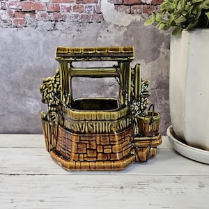 May include: A ceramic wishing well planter with a brown brick base and green accents. The words "Wishing Well" are inscribed on the front. A small bucket hangs from a chain on the side. Decorative item.