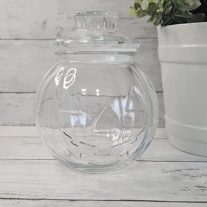 May include: A clear glass jar shaped like a pumpkin with a matching removable lid. The jar features a molded jack-o'-lantern face with triangular eyes, a triangular nose, and a wide, jagged grin embossed into the exterior surface of the glass.