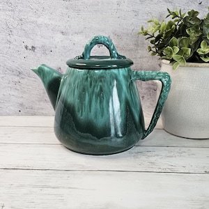 May include: A ceramic teapot featuring a glossy, deep emerald green glaze with darker forest green drips cascading down the sides. The teapot has a matching lid with a curved handle, a pointed spout, and a textured, braided-style side handle.