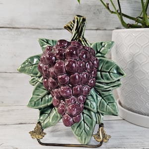 May include: A ceramic grapevine wall hanging with a cluster of purple grapes and green leaves. The hanging is on a gold metal stand.