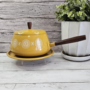 May include: Mustard yellow enamelware fondue pot with a matching lid and saucer. The pot features white daisy designs and a dark wood handle and lid knob. A vintage kitchenware item.