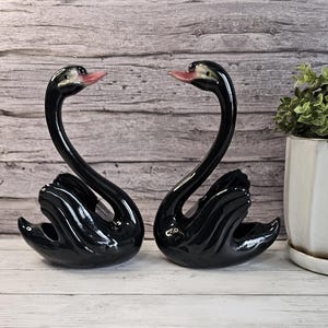 May include: A pair of glossy black ceramic swan figurines with pink beaks. The swans are facing each other, with their necks forming a heart shape. The figurines are displayed on a white surface.
