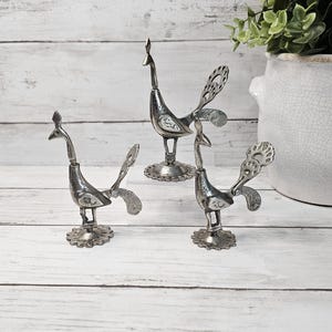 May include: Three silver-toned metal peacock figurines standing on circular, scalloped bases. Each bird features an elongated neck, a textured body, and a fanned tail with intricate cut-out patterns. The figures are crafted with a metallic finish.