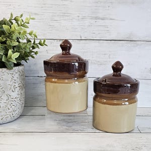 May include: Two antique ceramic jars with lids. The jars are two-toned, with a light tan base and a dark brown top and lid. The lids have a small, rounded knob. The jars are displayed on a clear stand against a white wood background.