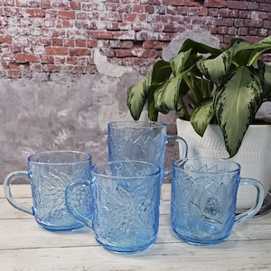 May include: Four light blue glass mugs with a floral design. The mugs have handles and are sitting on a wooden surface.