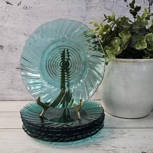 May include: A stack of five translucent teal glass plates featuring a central circular ripple pattern and a spiraling, fluted edge design. One plate is displayed upright on a gold-toned metal stand, while the remaining four plates are stacked.
