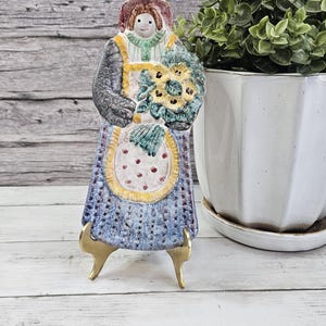 May include: A ceramic figurine of a woman holding a bouquet of yellow flowers. The figurine has a blue dress, a yellow apron with red dots, and a brown hat. It stands on gold-colored legs. A white potted plant is in the background.