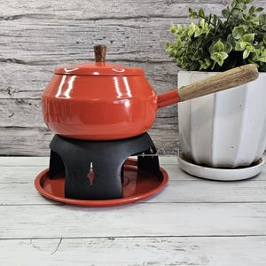 May include: A red fondue pot with a wooden handle and lid, sitting on a black stand and red base. The pot has a wooden knob on the lid. A white pot with greenery is to the right.