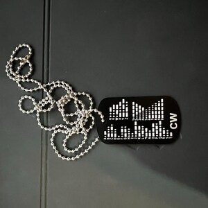 May include: Black dog tag pendant with a silver ball chain. The tag features a graphic design with the letters "CW" and a series of vertical lines and dots. The pendant is approximately 1.5 inches wide and 1 inch tall.
