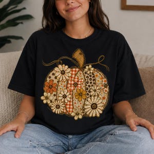 May include: A black short-sleeve t-shirt featuring a large graphic of a pumpkin. The pumpkin design is composed of various vertical panels with patterns including orange and white gingham, leopard print, and floral motifs in shades of yellow.