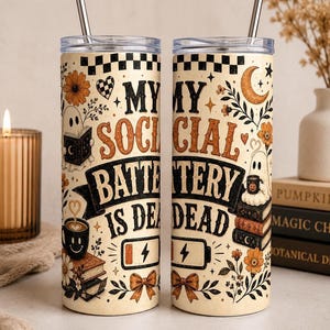 May include: Two cylindrical tumblers with clear lids and metal straws, featuring a cream background with black and orange illustrations of ghosts, books, flowers, and a battery icon. The text reads, "MY SOCIAL BATTERY IS DEAD" in bold lettering.