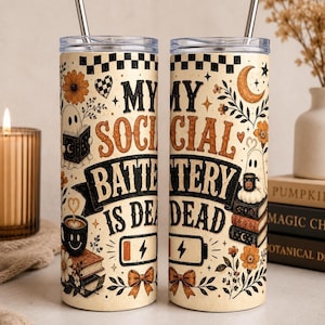 May include: Two cylindrical tumblers with clear lids and metal straws, featuring a cream background with black and orange illustrations of ghosts, books, flowers, bows, and a battery icon with the text MY SOCIAL BATTERY IS DEAD.