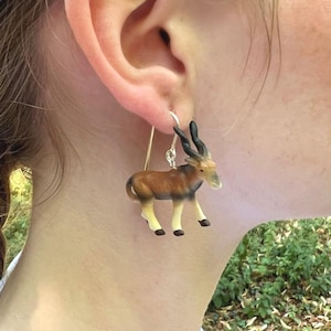 Op de afbeelding: Een gedetailleerde oorbel met een miniatuur antilope figuur. De antilope is bruin, beige en zwart, met gebogen hoorns. De oorbel hangt aan een goudkleurige draad haak, met een uniek dier-thema sieraad.