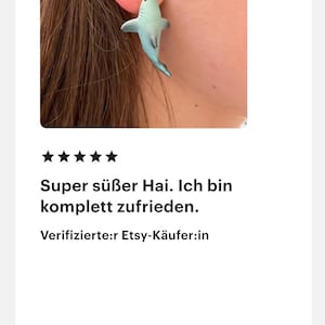 K&ouml;nnte beinhalten: Ein hellblauer Ohrring in Form eines Hais wird an einem Ohr getragen. Der Ohrring ist eine kleine, detaillierte Darstellung eines Hais. Der Text auf dem Bild lautet: "Super s&uuml;&szlig;er Hai. Ich bin komplett zufrieden."