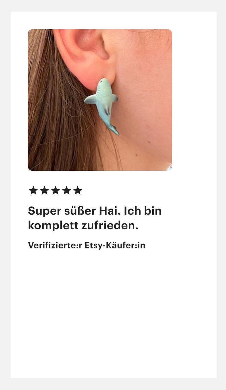 K&ouml;nnte beinhalten: Ein hellblauer Ohrring in Form eines Hais wird an einem Ohr getragen. Der Ohrring ist eine kleine, detaillierte Darstellung eines Hais. Der Text auf dem Bild lautet: "Super s&uuml;&szlig;er Hai. Ich bin komplett zufrieden."