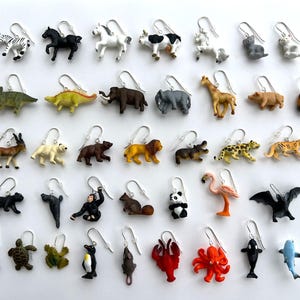 K&ouml;nnte beinhalten: Ohrringe mit Tiermotiven, darunter Zebra, Pferd, Kuh, Giraffe, B&auml;r, L&ouml;we und mehr. Jeder Ohrring hat einen kleinen Tieranh&auml;nger mit einem silberfarbenen Haken. Auf wei&szlig;em Hintergrund pr&auml;sentiert.
