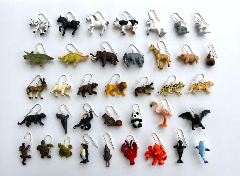 K&ouml;nnte beinhalten: Ohrringe mit Tiermotiven, darunter Zebra, Pferd, Kuh, Giraffe, B&auml;r, L&ouml;we und mehr. Jeder Ohrring hat einen kleinen Tieranh&auml;nger mit einem silberfarbenen Haken. Auf wei&szlig;em Hintergrund pr&auml;sentiert.