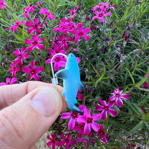 K&ouml;nnte beinhalten: Ein hellblauer Hai-Ohrring mit einem silbernen Haken wird vor einem Hintergrund aus leuchtend rosa Bl&uuml;ten und gr&uuml;nem Laub gehalten. Der Ohrring ist ein kleiner, detaillierter Anh&auml;nger.