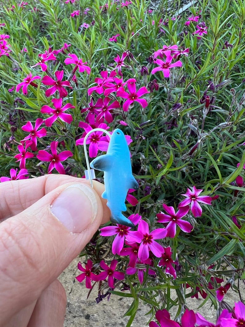 K&ouml;nnte beinhalten: Ein hellblauer Hai-Ohrring mit einem silbernen Haken wird vor einem Hintergrund aus leuchtend rosa Bl&uuml;ten und gr&uuml;nem Laub gehalten. Der Ohrring ist ein kleiner, detaillierter Anh&auml;nger.