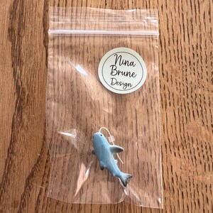 K&ouml;nnte beinhalten: Eine kleine, hellblaue Hai-Figur mit schwarzen Akzenten, verpackt in einer transparenten Plastikt&uuml;te. Der Hai hat einen wei&szlig;en Faden befestigt. Ein runder Aufkleber mit dem Text "Nina Brune Design" ist sichtbar.