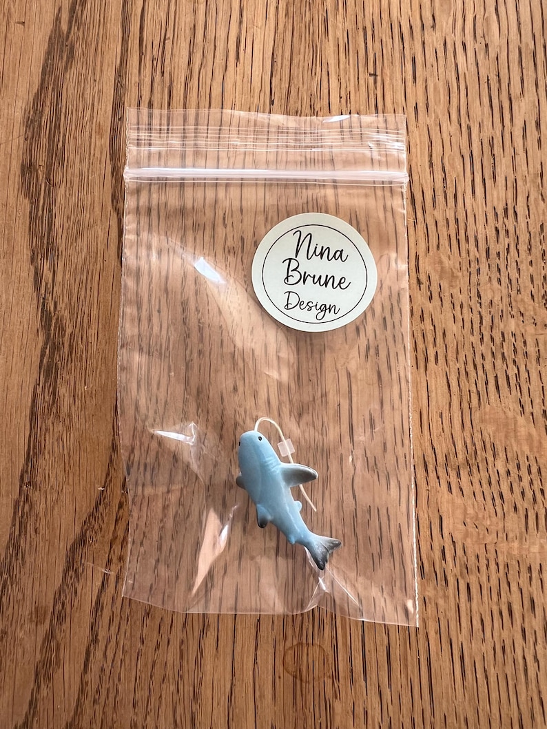 K&ouml;nnte beinhalten: Eine kleine, hellblaue Hai-Figur mit schwarzen Akzenten, verpackt in einer transparenten Plastikt&uuml;te. Der Hai hat einen wei&szlig;en Faden befestigt. Ein runder Aufkleber mit dem Text "Nina Brune Design" ist sichtbar.