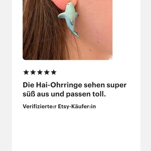 K&ouml;nnte beinhalten: Ein hellblauer Hai-Ohrring wird an einem Ohr getragen. Der Ohrring ist eine kleine, detaillierte Skulptur eines Hais. Der Text im Bild lautet: "Die Hai-Ohrringe sehen super s&uuml;&szlig; aus und passen toll."