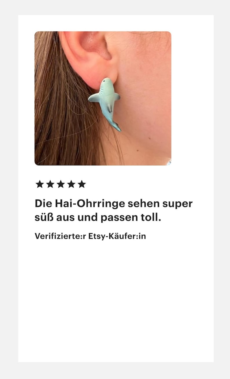 K&ouml;nnte beinhalten: Ein hellblauer Hai-Ohrring wird an einem Ohr getragen. Der Ohrring ist eine kleine, detaillierte Skulptur eines Hais. Der Text im Bild lautet: "Die Hai-Ohrringe sehen super s&uuml;&szlig; aus und passen toll."