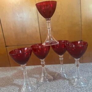 May include: A pyramid of five red wine glasses. The glasses have a clear stem and a ruby-red bowl. The glasses are arranged with four glasses at the base and one balanced on top.