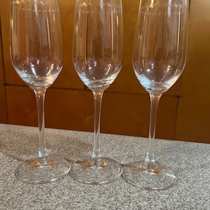 May include: Three clear champagne flutes with long stems and round bases. Each glass has a series of vertical lines and a dotted design near the rim. The glasses are arranged on a speckled gray surface.