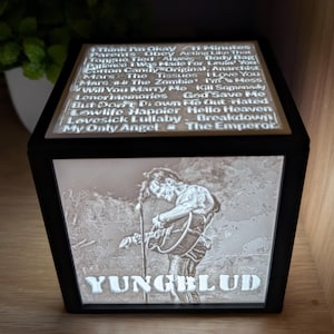May include: Black cube lamp with an engraved image of a musician playing guitar, the name "YUNGBLUD" below. The top surface lists song titles. The lamp is lit, emitting a warm light. The design is detailed and artistic.