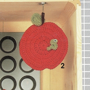 May include: A red crocheted apple with a green leaf and a green crocheted worm hanging from a hook. The apple is hanging over a muffin tin.