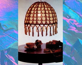Puede incluir: Una pantalla de lámpara de crochet con un patrón marrón y blanco y flecos. La pantalla está colgada del techo sobre una mesa con una taza y un platillo, un jarrón con flores y una cesta de mimbre.