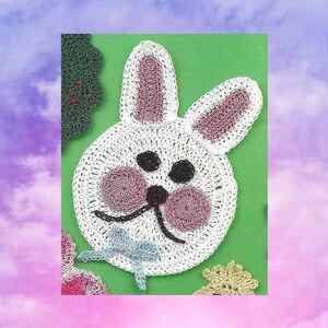 Könnte beinhalten: Ein weißer gehäkelter Hase mit rosa Ohren und einer blauen Schleife. Der Hase hat schwarze Augen und eine schwarze Nase. Der Hase befindet sich auf einem grünen Hintergrund.