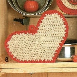 May include: A crocheted heart-shaped pot holder with a red border. The pot holder is made of beige yarn and is sitting on a wooden shelf.