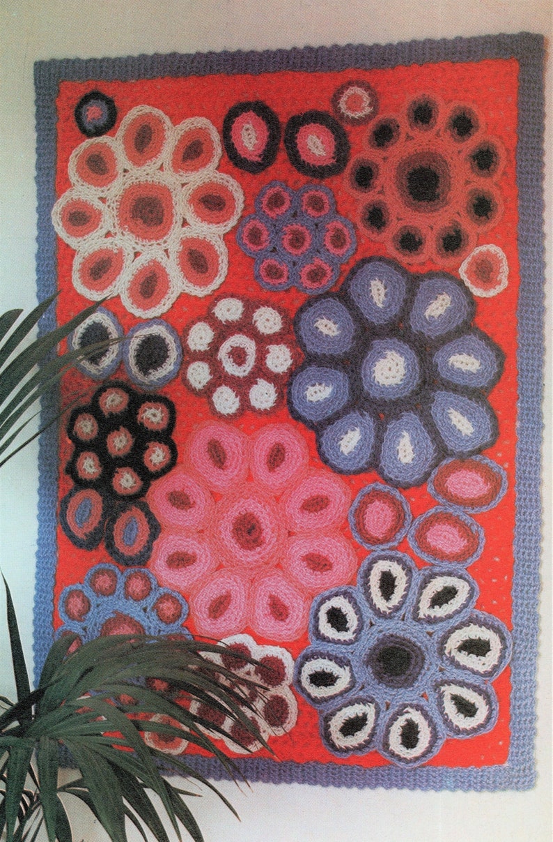 A 70's vintage, retro crochet rug hanging on a wall with a green plants nearby. The rug is bright pink, periwinkle blue and purple, white, baby blue, bark blue and magenta flowers.