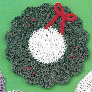 Puede incluir: Una corona de Navidad de ganchillo verde con un adorno de ganchillo blanco en el centro. Un lazo de ganchillo rojo está sujeto a la parte superior de la corona.