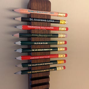 May include: A wooden pencil holder with 10 pencils, each with a different golf course name printed on it. The pencils are arranged vertically in the holder, with the eraser ends facing up. The holder is made of dark wood and has a grooved design.