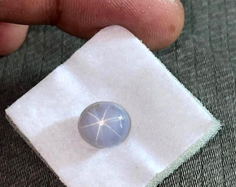 Könnte beinhalten: Ein runder, hellblauer Edelstein mit einem sechsstrahligen Sternmuster. Der Edelstein liegt auf einer weißen, quadratischen, strukturierten Oberfläche. Der Edelstein ist ein Sternsaphir, eine Art Korund. Der Hintergrund ist ein grauer Stoff.