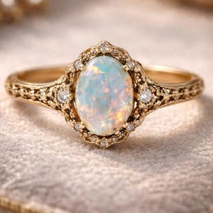Könnte beinhalten: Ein goldener Ring mit einem ovalen Opal-Edelstein. Der Opal ist von kleinen Diamanten umgeben und in einer dekorativen Fassung im Filigranstil gefasst. Das Band des Rings weist ebenfalls ein aufwendiges Design auf.