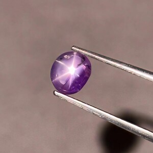 Pode incluir: Uma gema de safira estrela roxa com uma estrela de seis pontas no centro, segurada por pinças.