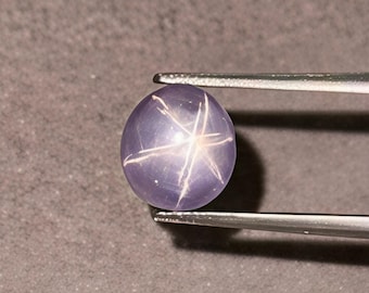 Könnte beinhalten: Ein lavendelfarbener Sternsaphir-Edelstein, der von einer Pinzette gehalten wird. Der ovale Edelstein hat ein sechsstrahliges Sternmuster auf seiner Oberfläche. Der polierte Edelstein hat eine hellviolette Farbe. Der Hintergrund ist ein neutrales Grau.