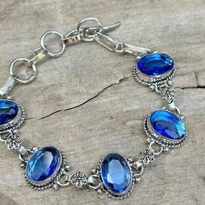 May include: A silver bracelet with five oval-shaped blue gemstones. The bracelet has a delicate chain and floral accents.