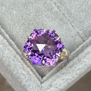 Puede incluir: Un anillo de plata con una gran piedra preciosa de amatista facetada. La piedra preciosa es de color morado oscuro y tiene un diseño de estrella.