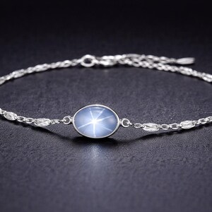 Könnte beinhalten: Ein silbernes Armband mit einem ovalen, blauen Edelstein in der Mitte, der ein sternförmiges Muster aufweist. Das Armband hat ein filigranes Kettendesign mit kleinen, dekorativen Gliedern. Der Edelstein ist in einer silbernen Lünette gefasst.