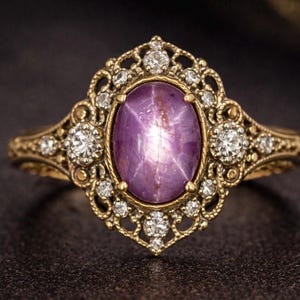 May include: A vintage-style ring with a gold band and a large, oval, purple gemstone. The gemstone has a star-like pattern. The ring is adorned with small, clear gemstones and intricate filigree detailing around the main stone.