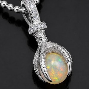 May include: A silver-toned pendant featuring a textured bird claw clutching a round, iridescent opal gemstone. The bail is embellished with small, clear, sparkling stones, and the piece hangs from a silver-toned ball chain necklace.