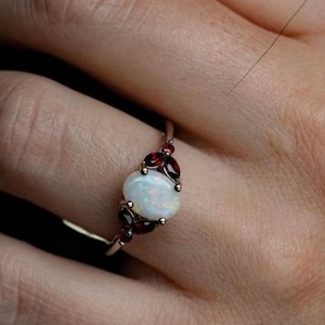 May include: A silver ring with an oval white opal gemstone surrounded by small red garnet stones.