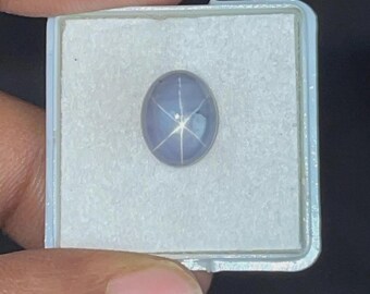 Könnte beinhalten: Ein hellblauer, ovaler Edelstein mit einem sechsstrahligen Sternmuster, in einem weißen Kunststoffgehäuse. Der Edelstein hat eine polierte, reflektierende Oberfläche. Der Sterneneffekt entsteht durch Lichtreflexionen.
